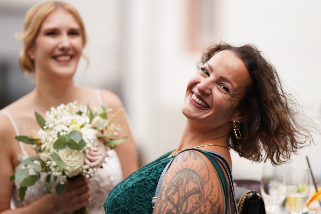 Hochzeitsfotografie einer jungen attraktiven Frau im Rahmen einer Hochzeit als Braut. Die Sonne scheint ihr ins Gesicht w&auml;hrend sie aus der Kirche von Hanau in der N&auml;he von Frankfurt am Main kommt. Sie tr&auml;gt ein schickes Brautkleid und sie h&auml;lt den Brautstrau&szlig; in der Hand. Neben ihr steht ihre Trauzeugin. Sie haben beide viel Spa&szlig; und lachen in die Kamera