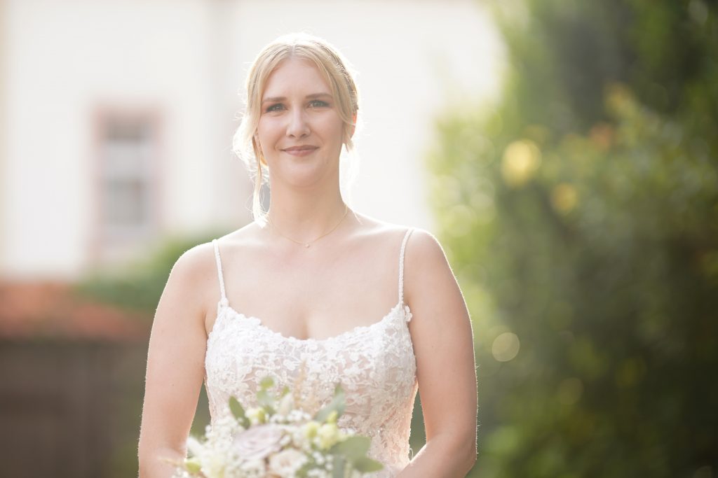 Hochzeitsfotografie einer jungen attraktiven Frau im Rahmen einer Hochzeit als Braut. Die Sonne scheint ihr ins Gesicht w&auml;hrend sie aus der Kirche von Hanau in der N&auml;he von Frankfurt am Main kommt. Sie tr&auml;gt ein schickes Brautkleid und sie h&auml;lt den Brautstrau&szlig; in der Hand.