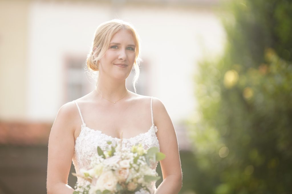 Hochzeitsfotografie einer jungen attraktiven Frau im Rahmen einer Hochzeit als Braut. Die Sonne scheint ihr ins Gesicht w&auml;hrend sie aus der Kirche von Hanau in der N&auml;he von Frankfurt am Main kommt. Sie tr&auml;gt ein schickes Brautkleid und sie h&auml;lt den Brautstrau&szlig; in der Hand.