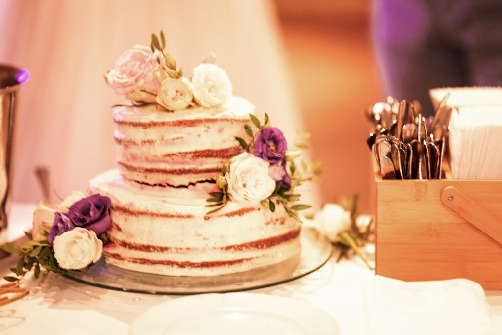 Hochzeitsfotografie vom Anschneiden der Hochzeitstorte eines Brautpaars in schickem Brautkleid und Anzug im Rahmen einer Hochzeit. Sie feiern in einer sch&ouml;nen Location in Hanau in der N&auml;he von Frankfurt am Main. Sie genie&szlig;en ihren besonderen Tag. Auf dem Hochzeitskuchen sind Blumen vom Brautstrau&szlig; als Deko platziert