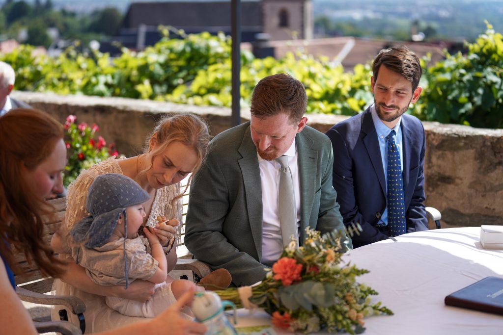 Hochzeitsfotografie von der standesamtlichen Trauung eines Brautpaars in schickem Brautkleid und Anzug im Rahmen einer Hochzeit. Die Sonne scheint in der Natur auf der Burg von Kronberg im Taunus in der N&auml;he von Frankfurt am Main. Sie tr&auml;gt ihren Brautstrau&szlig; in der Hand. Neben ihnen sitzen ihre Trauzeugen. Die Standesbeamte sitzt vor ihnen. Sie haben einen sch&ouml;nen Ausblick auf die Altstadt und den Taunus. Hinter ihnen sitzen ihre Familien.