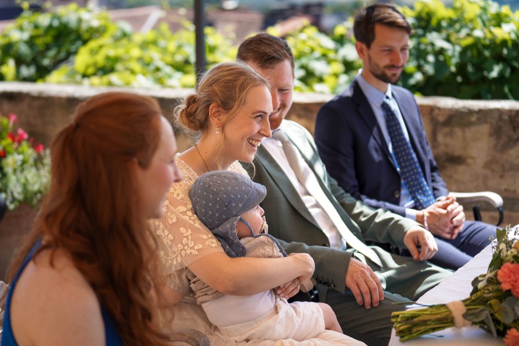 Hochzeitsfotografie von der standesamtlichen Trauung eines Brautpaars in schickem Brautkleid und Anzug im Rahmen einer Hochzeit. Die Sonne scheint in der Natur auf der Burg von Kronberg im Taunus in der N&auml;he von Frankfurt am Main. Sie tr&auml;gt ihren Brautstrau&szlig; in der Hand. Neben ihnen sitzen ihre Trauzeugen. Die Standesbeamte sitzt vor ihnen. Sie haben einen sch&ouml;nen Ausblick auf die Altstadt und den Taunus. Hinter ihnen sitzen ihre Familien.