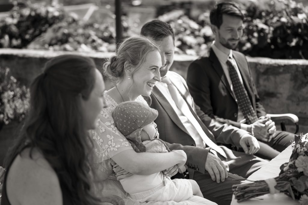 Schwarzwei&szlig; Hochzeitsfotografie von der standesamtlichen Trauung eines Brautpaars in schickem Brautkleid und Anzug im Rahmen einer Hochzeit. Die Sonne scheint in der Natur auf der Burg von Kronberg im Taunus in der N&auml;he von Frankfurt am Main. Sie tr&auml;gt ihren Brautstrau&szlig; in der Hand. Neben ihnen sitzen ihre Trauzeugen. Die Standesbeamte sitzt vor ihnen. Sie haben einen sch&ouml;nen Ausblick auf die Altstadt und den Taunus. Hinter ihnen sitzen ihre Familien.