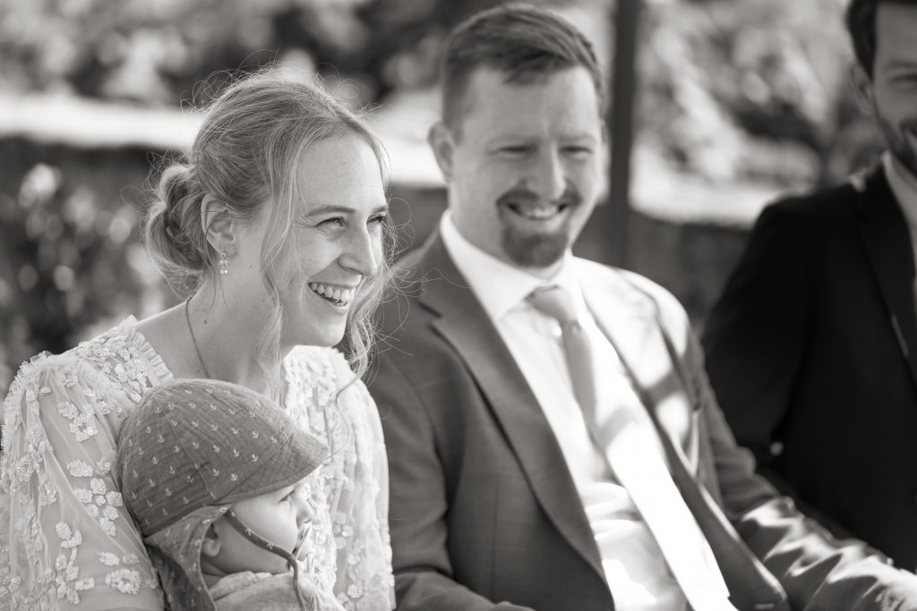 Schwarzwei&szlig; Hochzeitsfotografie von der standesamtlichen Trauung eines Brautpaars in schickem Brautkleid und Anzug im Rahmen einer Hochzeit. Die Sonne scheint in der Natur auf der Burg von Kronberg im Taunus in der N&auml;he von Frankfurt am Main. Sie tr&auml;gt ihren Brautstrau&szlig; in der Hand. Neben ihnen sitzen ihre Trauzeugen. Die Standesbeamte sitzt vor ihnen. Sie haben einen sch&ouml;nen Ausblick auf die Altstadt und den Taunus. Hinter ihnen sitzen ihre Familien.