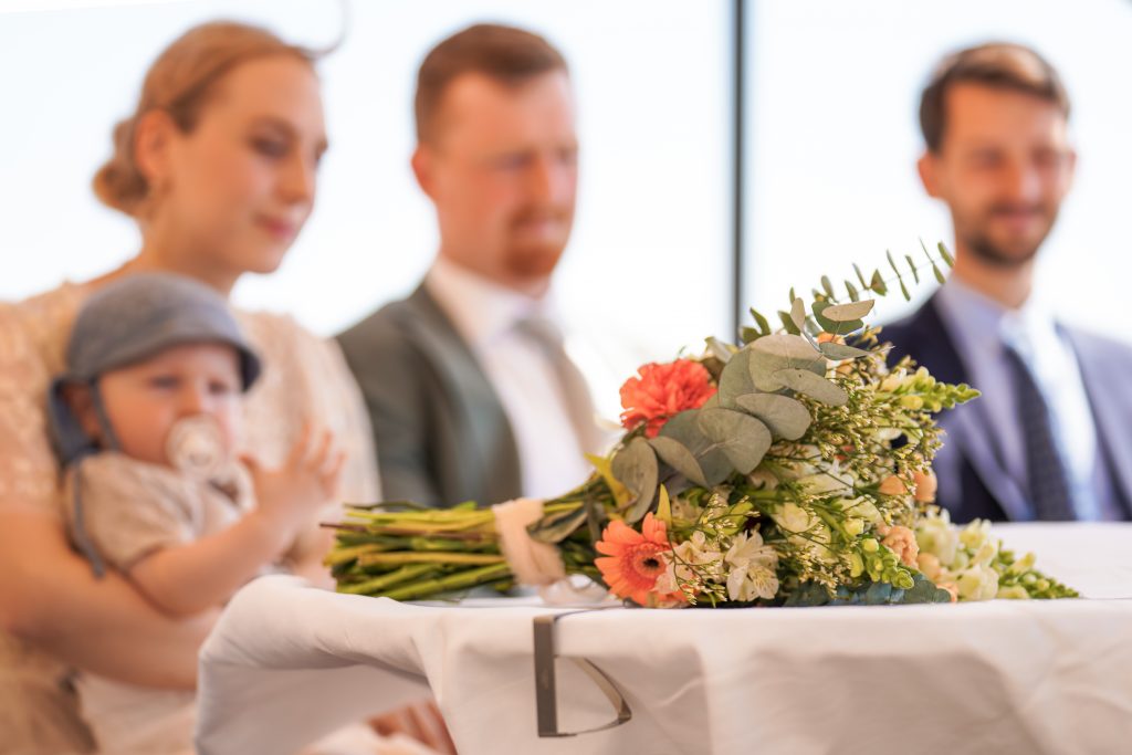 Hochzeitsfotografie von der standesamtlichen Trauung eines Brautpaars in schickem Brautkleid und Anzug im Rahmen einer Hochzeit. Die Sonne scheint in der Natur auf der Burg von Kronberg im Taunus in der N&auml;he von Frankfurt am Main. Sie tr&auml;gt ihren Brautstrau&szlig; in der Hand. Neben ihnen sitzen ihre Trauzeugen. Die Standesbeamte sitzt vor ihnen. Sie haben einen sch&ouml;nen Ausblick auf die Altstadt und den Taunus. Hinter ihnen sitzen ihre Familien.