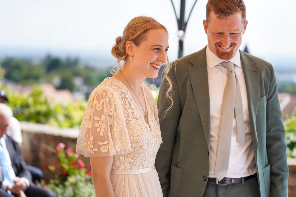 Hochzeitsfotografie von der standesamtlichen Trauung eines Brautpaars in schickem Brautkleid und Anzug im Rahmen einer Hochzeit. Die Sonne scheint in der Natur auf der Burg von Kronberg im Taunus in der N&auml;he von Frankfurt am Main. Sie tr&auml;gt ihren Brautstrau&szlig; in der Hand. Neben ihnen sitzen ihre Trauzeugen. Die Standesbeamte sitzt vor ihnen. Sie haben einen sch&ouml;nen Ausblick auf die Altstadt und den Taunus. Hinter ihnen sitzen ihre Familien.