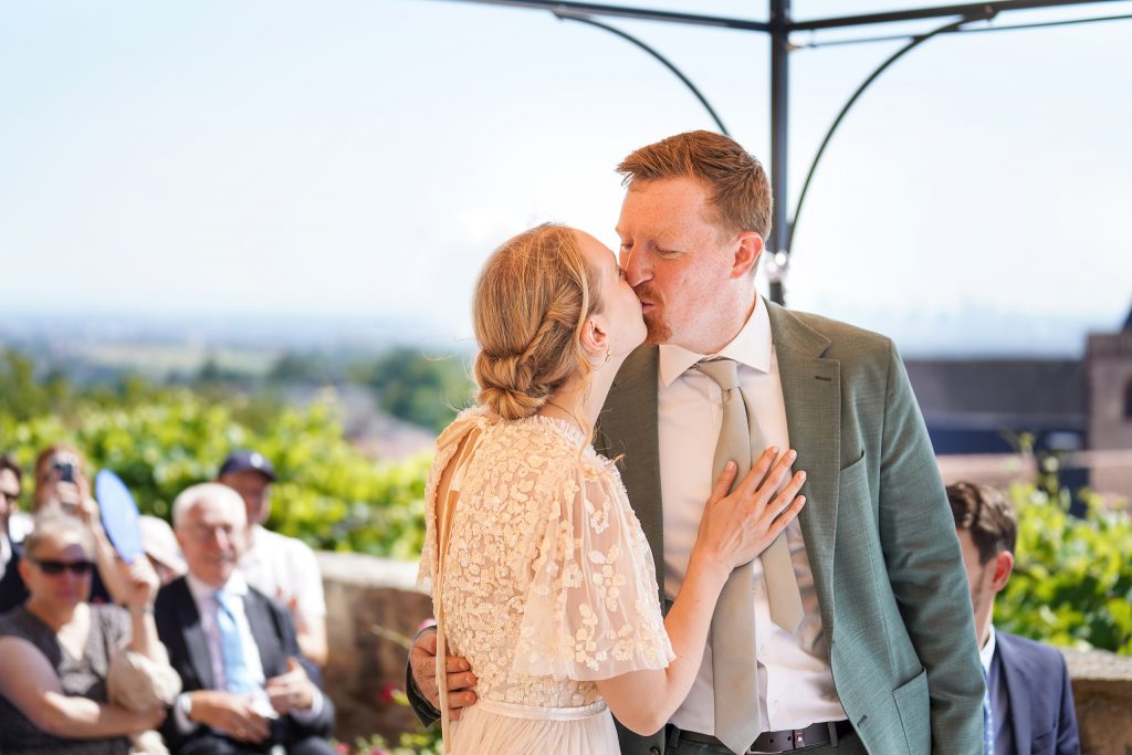 Hochzeitsfotografie von der standesamtlichen Trauung eines Brautpaars in schickem Brautkleid und Anzug im Rahmen einer Hochzeit. Die Sonne scheint in der Natur auf der Burg von Kronberg im Taunus in der N&auml;he von Frankfurt am Main. Sie tr&auml;gt ihren Brautstrau&szlig; in der Hand. Neben ihnen sitzen ihre Trauzeugen. Die Standesbeamte sitzt vor ihnen. Sie haben einen sch&ouml;nen Ausblick auf die Altstadt und den Taunus. Hinter ihnen sitzen ihre Familien. Sie k&uuml;ssen sich.