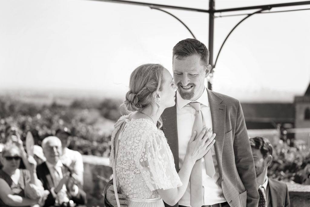 Schwarzwei&szlig; Hochzeitsfotografie von der standesamtlichen Trauung eines Brautpaars in schickem Brautkleid und Anzug im Rahmen einer Hochzeit. Die Sonne scheint in der Natur auf der Burg von Kronberg im Taunus in der N&auml;he von Frankfurt am Main. Sie tr&auml;gt ihren Brautstrau&szlig; in der Hand. Neben ihnen sitzen ihre Trauzeugen. Die Standesbeamte sitzt vor ihnen. Sie haben einen sch&ouml;nen Ausblick auf die Altstadt und den Taunus. Hinter ihnen sitzen ihre Familien. Sie k&uuml;ssen sich.