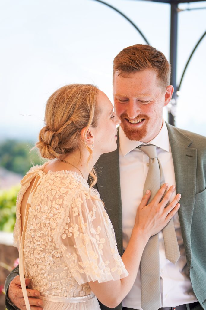Hochzeitsfotografie von der standesamtlichen Trauung eines Brautpaars in schickem Brautkleid und Anzug im Rahmen einer Hochzeit. Die Sonne scheint in der Natur auf der Burg von Kronberg im Taunus in der N&auml;he von Frankfurt am Main. Sie tr&auml;gt ihren Brautstrau&szlig; in der Hand. Neben ihnen sitzen ihre Trauzeugen. Die Standesbeamte sitzt vor ihnen. Sie haben einen sch&ouml;nen Ausblick auf die Altstadt und den Taunus. Hinter ihnen sitzen ihre Familien. Sie k&uuml;ssen sich.