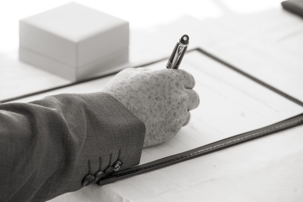 Schwarzwei&szlig; Hochzeitsfotografie von der standesamtlichen Trauung eines Brautpaars in schickem Brautkleid und Anzug im Rahmen einer Hochzeit. Die Sonne scheint in der Natur auf der Burg von Kronberg im Taunus in der N&auml;he von Frankfurt am Main. Sie steckt ihrem Mann den Ehering an die Hand und der steckt ihr den Ring an ihre Hand. Neben ihnen sitzen ihre Trauzeugen. Die Standesbeamte sitzt vor ihnen. Sie haben einen sch&ouml;nen Ausblick auf die Altstadt und den Taunus. Hinter ihnen sitzen ihre Familien.