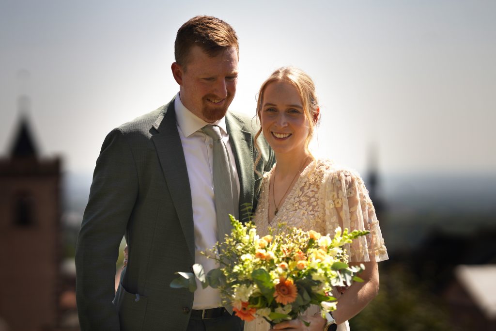 P&auml;rchenshooting und Hochzeitsfotografie vom Kuss eines Brautpaars in schickem Brautkleid und Anzug im Rahmen einer Hochzeit. Die Sonne scheint ihr ins Gesicht w&auml;hrend sie in der Natur der Burg von Kronberg im Taunus in der N&auml;he von Frankfurt am Main stehen. Sie tr&auml;gt ihren Brautstrau&szlig; in der Hand