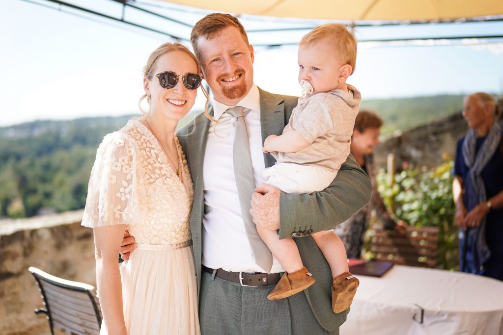 Hochzeitsfotografie von der standesamtlichen Trauung eines Brautpaars in schickem Brautkleid und Anzug im Rahmen einer Hochzeit. Die Sonne scheint in der Natur auf der Burg von Kronberg im Taunus in der N&auml;he von Frankfurt am Main. Sie steckt ihrem Mann den Ehering an die Hand und der steckt ihr den Ring an ihre Hand. Neben ihnen sitzen ihre Trauzeugen. Die Standesbeamte sitzt vor ihnen. Sie haben einen sch&ouml;nen Ausblick auf die Altstadt und den Taunus. Hinter ihnen sitzen ihre Familien.