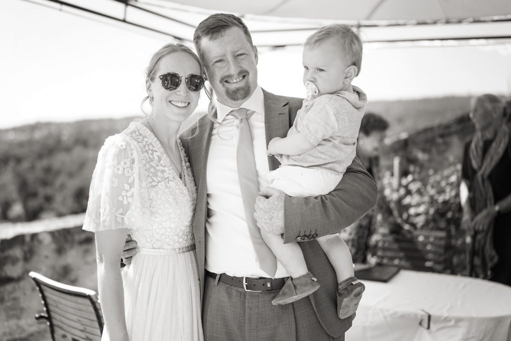 Schwarzwei&szlig; Hochzeitsfotografie von der standesamtlichen Trauung eines Brautpaars in schickem Brautkleid und Anzug im Rahmen einer Hochzeit. Die Sonne scheint in der Natur auf der Burg von Kronberg im Taunus in der N&auml;he von Frankfurt am Main. Sie tr&auml;gt ihren Brautstrau&szlig; in der Hand. Neben ihnen sitzen ihre Trauzeugen. Die Standesbeamte sitzt vor ihnen. Sie haben einen sch&ouml;nen Ausblick auf die Altstadt und den Taunus. Hinter ihnen sitzen ihre Familien.