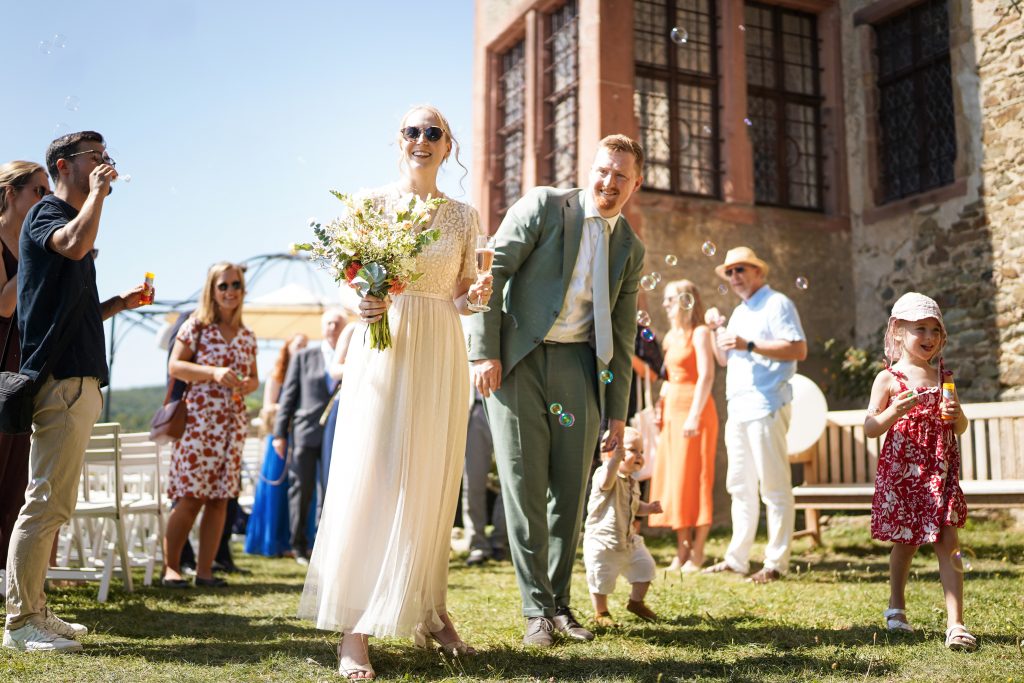 Hochzeitsfotografie von der standesamtlichen Trauung eines Brautpaars in schickem Brautkleid und Anzug im Rahmen einer Hochzeit. Die Sonne scheint in der Natur auf der Burg von Kronberg im Taunus in der N&auml;he von Frankfurt am Main. Sie steckt ihrem Mann den Ehering an die Hand und der steckt ihr den Ring an ihre Hand. Neben ihnen sitzen ihre Trauzeugen. Die Standesbeamte sitzt vor ihnen. Sie haben einen sch&ouml;nen Ausblick auf die Altstadt und den Taunus. Hinter ihnen sitzen ihre Familien.