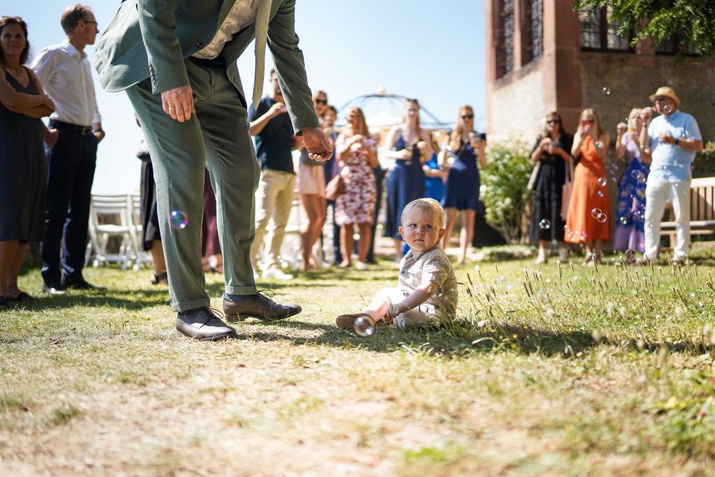 Hochzeitsfotografie von der standesamtlichen Trauung eines Brautpaars in schickem Brautkleid und Anzug im Rahmen einer Hochzeit. Die Sonne scheint in der Natur auf der Burg von Kronberg im Taunus in der N&auml;he von Frankfurt am Main. Sie steckt ihrem Mann den Ehering an die Hand und der steckt ihr den Ring an ihre Hand. Neben ihnen sitzen ihre Trauzeugen. Die Standesbeamte sitzt vor ihnen. Sie haben einen sch&ouml;nen Ausblick auf die Altstadt und den Taunus. Hinter ihnen sitzen ihre Familien.