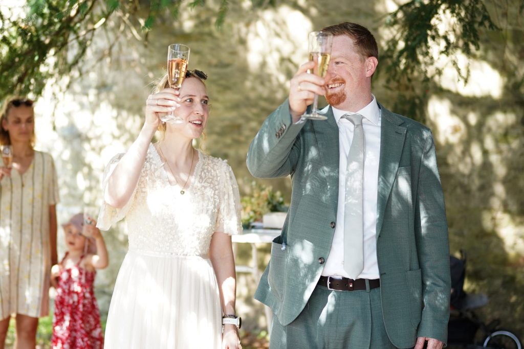 Hochzeitsfotografie von der standesamtlichen Trauung eines Brautpaars in schickem Brautkleid und Anzug im Rahmen einer Hochzeit. Die Sonne scheint in der Natur auf der Burg von Kronberg im Taunus in der N&auml;he von Frankfurt am Main. Sie steckt ihrem Mann den Ehering an die Hand und der steckt ihr den Ring an ihre Hand. Neben ihnen sitzen ihre Trauzeugen. Die Standesbeamte sitzt vor ihnen. Sie haben einen sch&ouml;nen Ausblick auf die Altstadt und den Taunus. Hinter ihnen sitzen ihre Familien. Sie haben viel Spa&szlig; auf ihrer Hochzeitsfeier und Party.