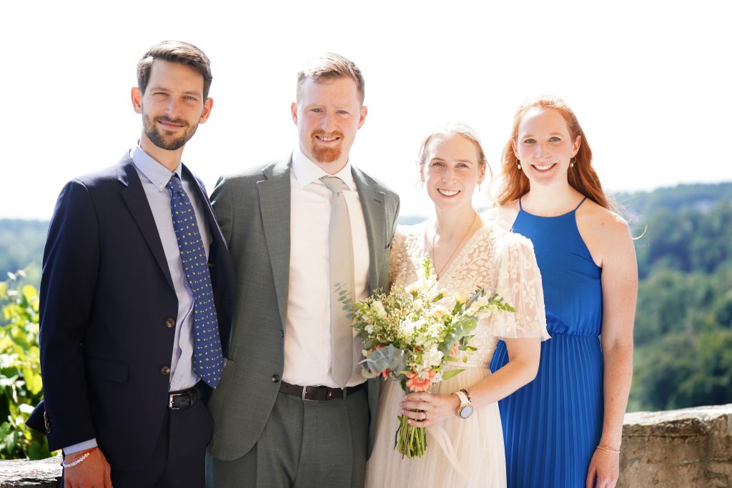 Hochzeitsfotografie von der standesamtlichen Trauung eines Brautpaars in schickem Brautkleid und Anzug im Rahmen einer Hochzeit. Die Sonne scheint in der Natur auf der Burg von Kronberg im Taunus in der N&auml;he von Frankfurt am Main. Sie steckt ihrem Mann den Ehering an die Hand und der steckt ihr den Ring an ihre Hand. Neben ihnen sitzen ihre Trauzeugen. Die Standesbeamte sitzt vor ihnen. Sie haben einen sch&ouml;nen Ausblick auf die Altstadt und den Taunus. Hinter ihnen sitzen ihre Familien. Sie haben viel Spa&szlig; auf ihrer Hochzeitsfeier und Party.