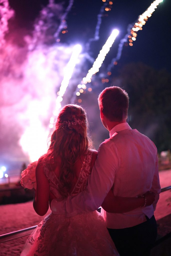 Hochzeitsfotografie vom Kuss eines Brautpaars in schickem Brautkleid und Anzug im Rahmen einer Hochzeit. Hinter ihnen leuchtet das Feuerwerk w&auml;hrend sie in der Natur in der N&auml;he von Frankfurt am Main stehen