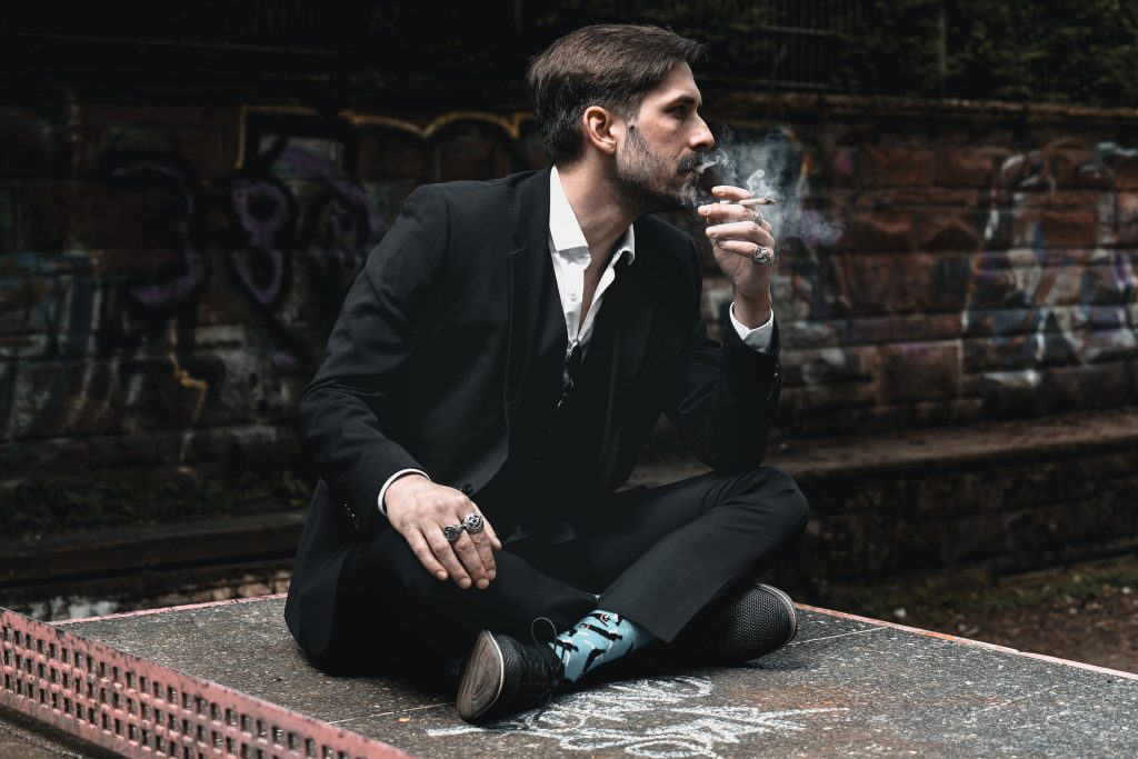 Streetphotography-Fashion-Fotografie-Anzug-Park-Frankfurt-Streetart-Grafitti-Rauch Street-Fotografie von jungen modischen Mann in modernem Anzug in der Frankfurter Innenstadt imeinem Park auf einer Tischtennisplatte beim rauchen. Der Rauch schwebt vor ihn dzrcg die Luft. Auf der Mauer im Hintergrund sind Graffiti zu sehen.