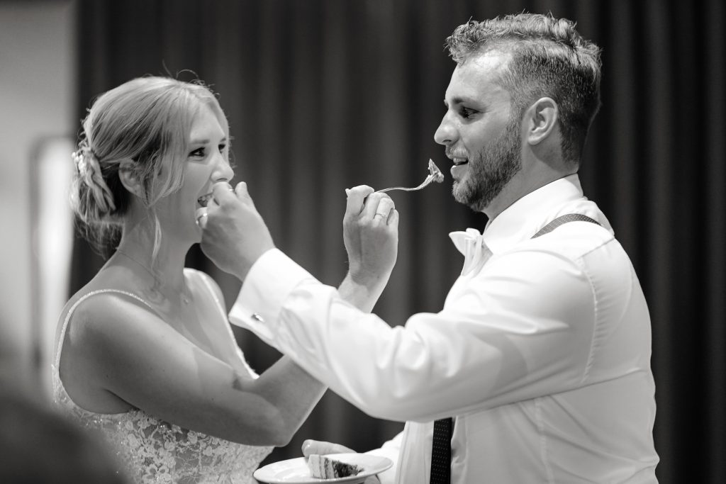 Schwarzweiß-Hochtzeitsfotografie-Braut-Bräutigam-glücklich-Kleid-Hochzeitstorte-Schön-Brautkleid-Hanau-Frankfurt-Standesamt-Kuss Schwarzweiß Hochzeitsfotografie vom Anschneiden der Hochzeitstorte eines Brautpaars in schickem Brautkleid und Anzug im Rahmen einer Hochzeit. Sie feiern in einer schönen Location in Hanau in der Nähe von Frankfurt am Main. Sie genießen ihren besonderen Tag