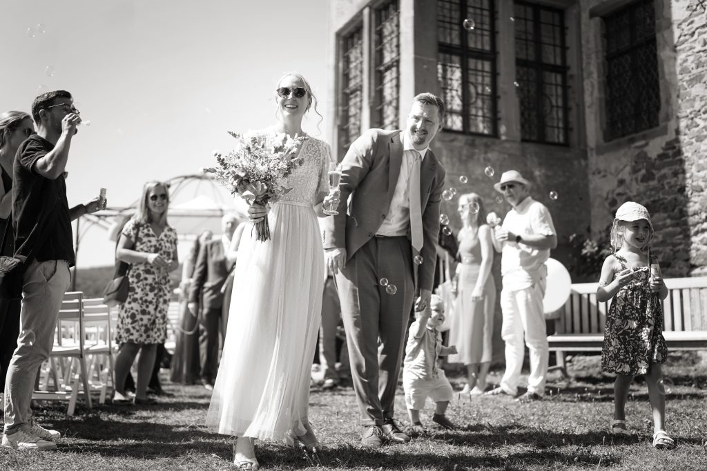 Schwarzweiß-Hochtzeitsfotografie-Braut-Bräutigam-glücklich-Kleid-Sonne-Schön-Brautkleid-Natur-Frankfurt-Standesamt-Kronberg-Taunus-Burg-Kuss-Brautstrauß-Trauung Schwarzweiß Hochzeitsfotografie vom Brautpaar in schickem Brautkleid und Anzug im Rahmen einer Hochzeit. Die Sonne scheint ihr ins Gesicht während sie in der Natur der Burg von Kronberg im Taunus in der Nähe von Frankfurt am Main stehen. Sie trägt ihren Brautstrauß in der Hand und sie genießen den Moment nach ihrer Trauung im Rahmen ihrer Familie und ihren Freunden