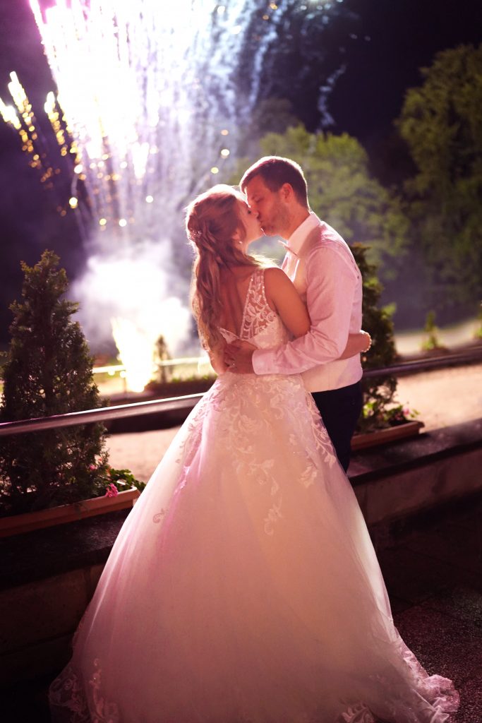 Hochtzeitsfotografie-Braut-Bräutigam-glücklich-Kleid-Feuerwerk-Traumhochzeit-Brautkleid-Natur-Frankfurt-Kuss-Brautstrauß Hochzeitsfotografie vom Kuss eines Brautpaars in schickem Brautkleid und Anzug im Rahmen einer Hochzeit. Hinter ihnen leuchtet das Feuerwerk während sie in der Natur in der Nähe von Frankfurt am Main stehen