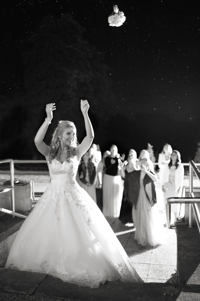 Schwarzweiß-Hochtzeitsfotografie-Braut-Bräutigam-glücklich-Kleid-Hochzeitstorte-Brautkleid-Hanau-Frankfurt-Brautstrauß-Braustraußwurf-Sterne-Trauzäugin Schwarzweiß Hochzeitsfotografie vom Wurf des Brautstraußes der Braut in schickem Brautkleid zu ihren Trauzeuginnen im Rahmen einer Hochzeit. Die Sterne funkeln im Himmel in der Nähe von Frankfurt am Main.
